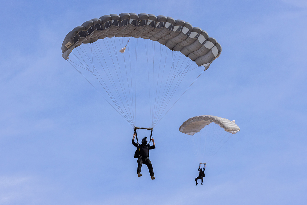 Two of the six Exercise Rising Gains 1‑26 parachutists about to land with open grey coloured rectangular parachutes, line astern.
